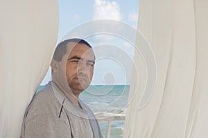 Portrait of a man at window with white curtains. Sea or ocean in the background