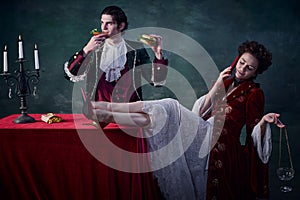 Portrait of man and woman in image of medieval vampires over dark green background. Man eating burger and drinking
