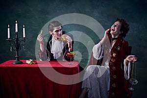 Portrait of man and woman in image of medieval vampires over dark green background. Man eating burger, woman talking on