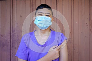 Portrait of a masked rural Asian boy looking at the camera with his hand pointing at plaster after being vaccinated against