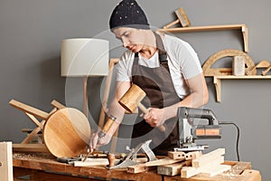 Portrait of master wearing brown apron and black cap using mallet and chisel to smooth rectangular block of wood while it stands