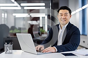 Portrait of mature asian businessman, successful experienced man with laptop inside office smiling and looking at camera