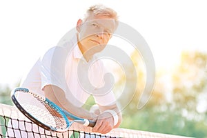 Portrait of mature athlete leaning on tennis net in court with yellow lens flare in background