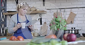 Portrait of mature blond woman drinking coffee and reading book of recipes in the morning. Serious middle-aged retiree