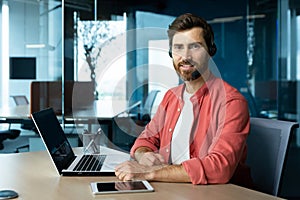 Portrait of mature businessman in red shirt inside office, man with beard and video call headset smiling and looking at