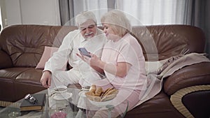 Portrait of mature caucasian man and woman using smartphone at home. Modern retirees sitting on couch indoors talking