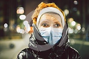 Portrait mature Caucasian woman in protective medical mask on face against background of evening city in winter. An elderly woman