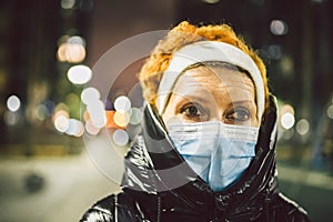 Portrait mature Caucasian woman in protective medical mask on face against background of evening city in winter. An elderly woman