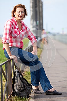 Portrait of mature Caucasian woman in red t-shirt and bluse jeans sitting with backpack on streets