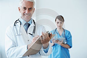Portrait, mature doctor writing on his clipboard and with a nurse browsing on a digital tablet in the background. Senior