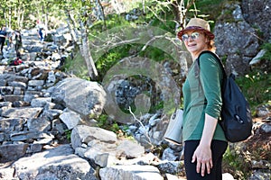Portrait of mature European woman hiker standing near stone way to mountain, backpack is on back