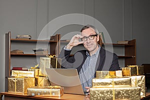 Portrait of mature excited businessman in glasses and blue suit sitting by the table with laptop and a lot of gifts around