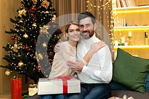 Portrait of mature family bearded man and blonde woman looking at camera hugging near Christmas tree, couple celebrating