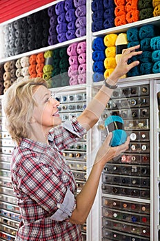 portrait of mature female customer standing next to shelf with knitting yarn in shop