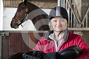 Portrait Of Mature Female Owner Holding Saddle In Stable With Ho