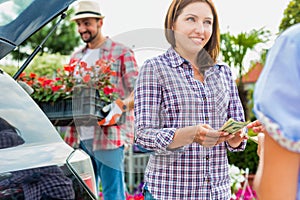 Portrait of mature gardener putting flowers on crate in car trunk while woman buyer giving cash payment