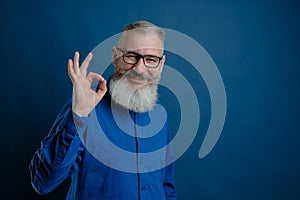 Portrait of mature gray-haired bearded caucasian man dressed blue shirt shows ok sign, blue background, selective focus