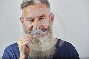 Portrait of mature gray-haired bearded man in blue sweatshirt with mustache comb on gray background, selective focus