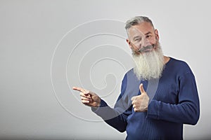 Portrait of mature gray-haired bearded man points forefinger to side and shows thumb up on gray background, selective focus