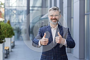 Portrait of mature investor outside office building, senior man smiling and looking at camera showing fingers thumbs up