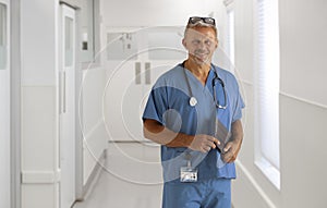 Portrait Of Mature Male Doctor Or Surgeon Wearing Scrubs In Hospital Corridor With Digital Tablet
