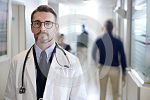 Portrait Of Mature Male Doctor Wearing White Coat With Stethoscope In Busy Hospital Corridor