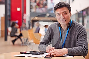 Portrait Of Mature Male Teacher Or Student With Digital Tablet Working At Table In College Hall