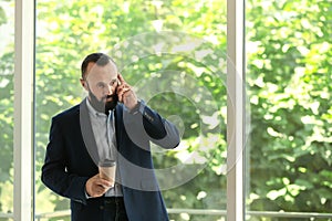 Portrait of mature man in elegant suit with coffee talking on mobile phone near window