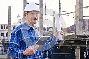 portrait mature man engineer in checkered shirt wears safety helmet, one hand holding tablet and other hand holding blueprint