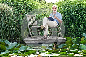 Portrait Of Mature Man Relaxing In Garden Reading Book On Jetty By Lake