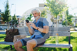 Portrait of mature man sitting outdoors on bench in city, using smartphone.