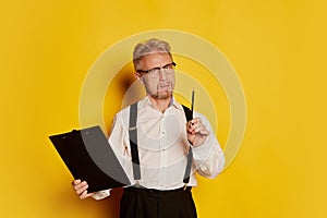 Portrait of mature man in stylish classical clothes posing with tablet and pencil isolated over yellow studio background