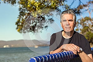 Portrait of a mature man 57 years with gray hair looking camera sitting on a bench at a beach outdoors, confident