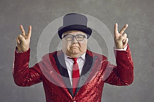 Portrait of funny serious rich senior man in top hat and sequin jacket showing V sign