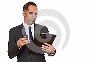 Portrait of mature Persian businessman using phone while reading on clipboard