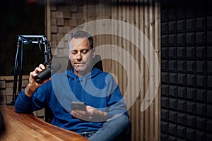 Portrait of mature radio host reading script in cellphone and speaking in microphone while moderating a live show