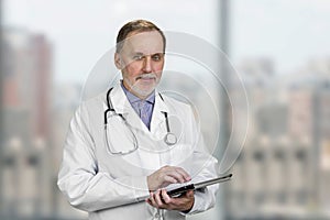 Portrait of mature senior male doctor holding digital tablet standing indoors.