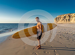 Portrait of mature senior Surfer looking at the ocean with vintage surfboard on an empty beach