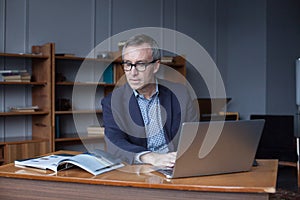 Portrait of mature serious businessman in suit and glasses working on the laptop and reading book simultaneously in office