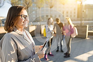 Portrait of mature smiling female teacher in glasses with clipboard, outdor with a group of teenagers students