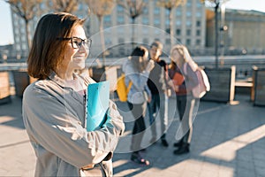 Portrait of mature smiling female teacher in glasses with clipboard, outdor with a group of teenagers students, golden hour