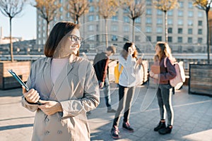 Portrait of mature smiling female teacher in glasses with clipboard, outdor with a group of teenagers students, golden hour
