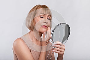 Portrait of mature woman checking her wrinkles in miror, looking at signs of aging on her face over light background