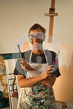 Portrait of mature woman wearing glasses with paint-stained face and crossed arms with brushes in her hands