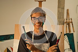 Portrait of mature woman wearing glasses with paint-stained face and crossed arms with brushes in her hands