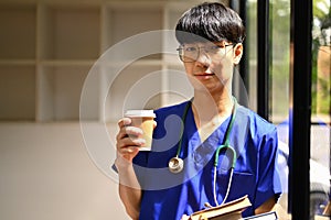 Portrait of medical student man wearing blue scrubs standing near window and looking at camera