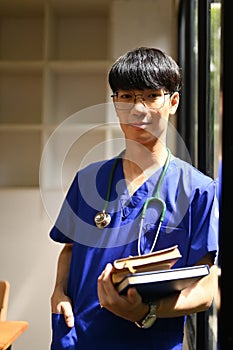 Portrait of medical student man wearing blue scrubs with stethoscope standing near window at campus