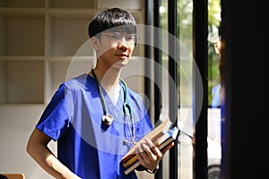 Portrait of medical student man wearing blue scrubs with stethoscope standing near window in sunny day in medical office
