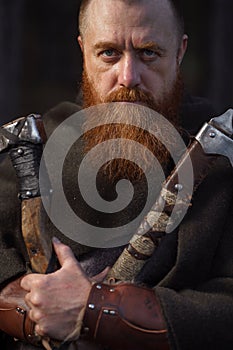 Portrait of medieval red-haired viking warrior with beard with two axes