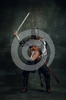 Portrait of medieval warrior or knight wearing helmet and armor riding toy horse, holding big sword  over dark